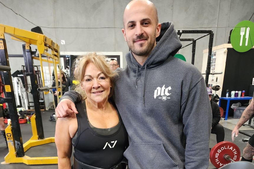 An older woman with blonde hair stands with arm around waist of taller man in hoodie in gym