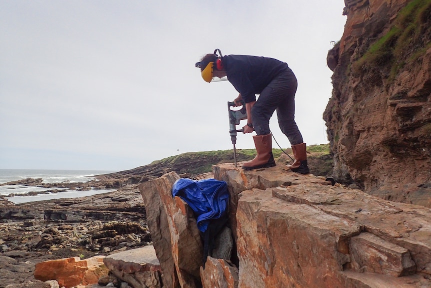 A woman in winter clothing and yellow visor drills stands on top of a rock she drills into by the sea.