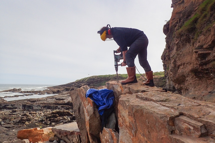 Une femme vêtue de vêtements d'hiver et d'exercices à visière jaune se dresse au sommet d'un rocher qu'elle perce au bord de la mer.