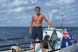 A sunburnt, man with grey beard stands shirtless on a boat with the Australian flag.