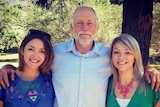 A man wearing a business shirt stands with his arms around two young women.
