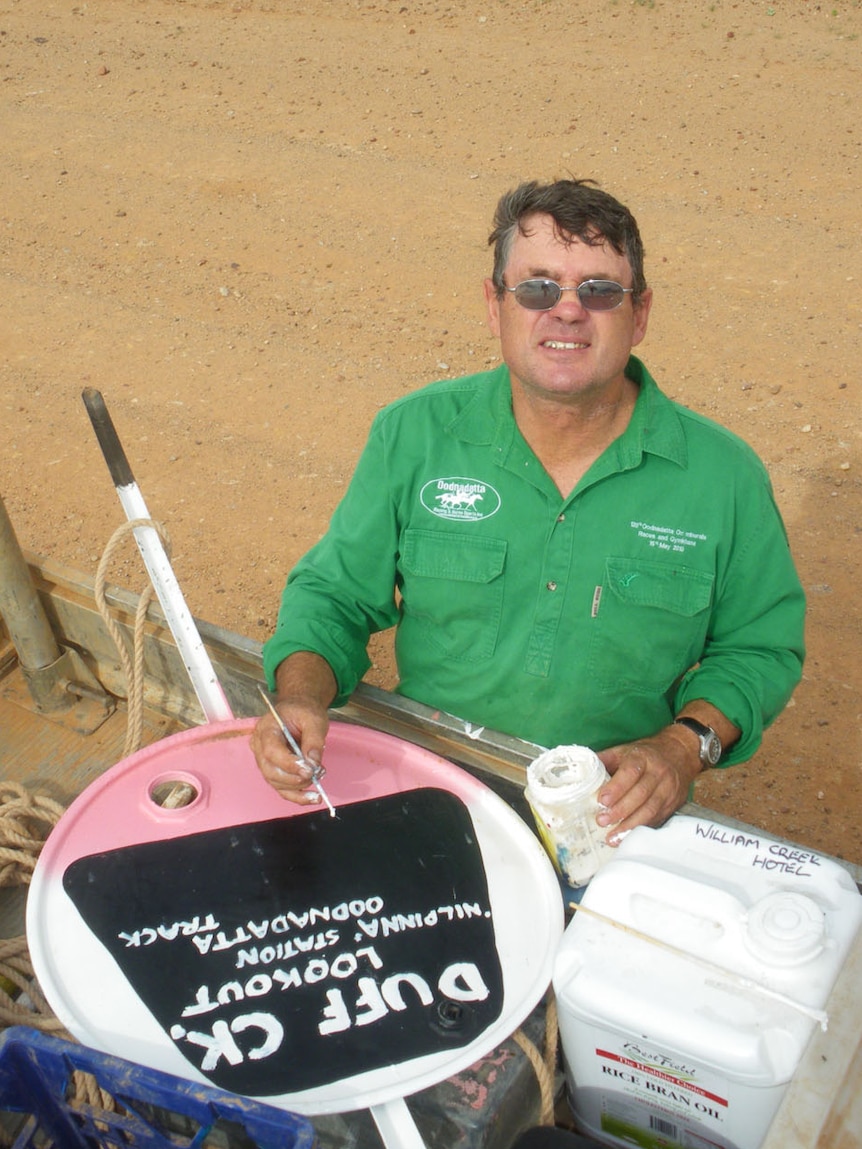 A man in a green shirt hand paints a road sign in the outback on the back of a ute.