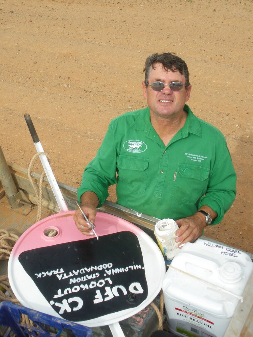 A man in a green shirt hand paints a road sign in the outback on the back of a ute.