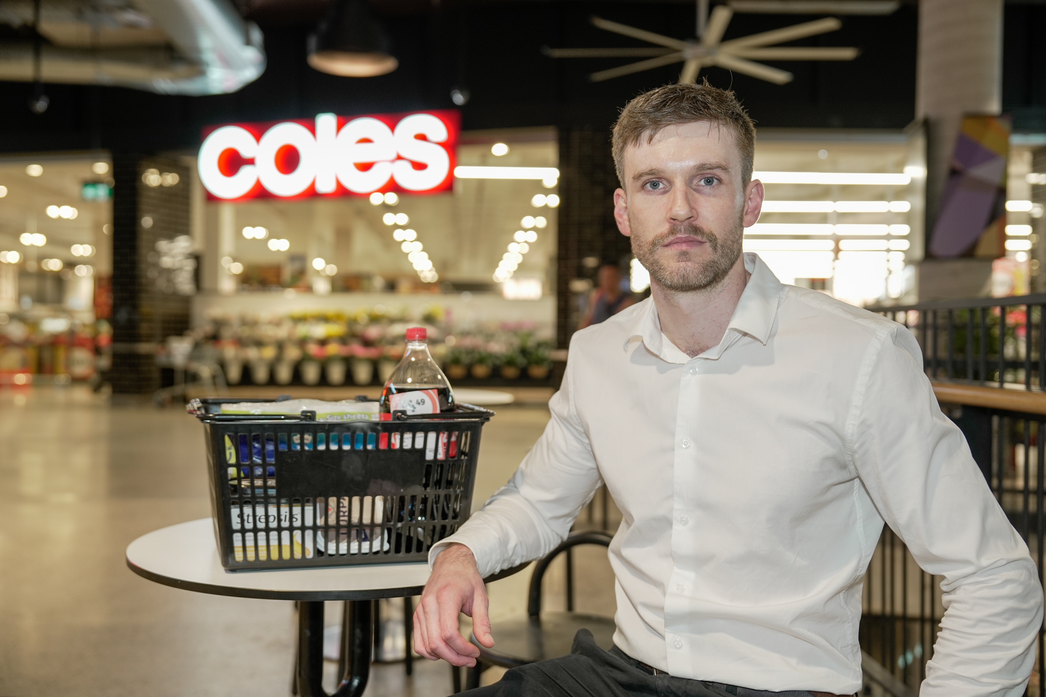 A man in a white button up shirt sits at a table and chairs in front of Coles with a shopping basket full of groceries.