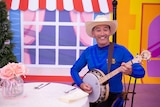 A man in a blue skivvy – Anthony Field, of The Wiggles – plays a banjo on what appears to be a TV show set.