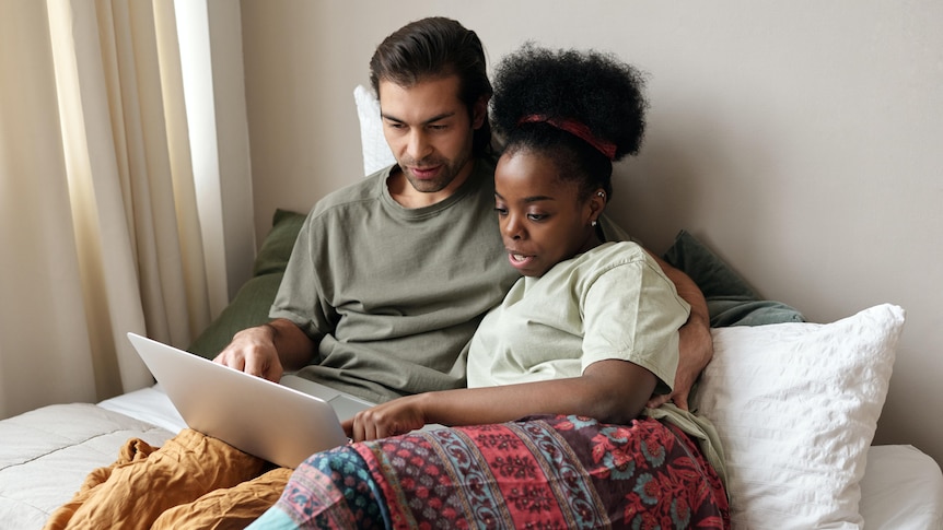 A man and a woman sitting up on a bed, speaking about something on their laptop.