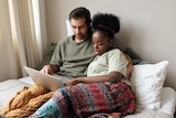 A man and a woman sitting up on a bed, speaking about something on their laptop.