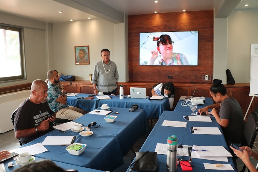 People sit around a table and look at a big projector screen where a women is taking.