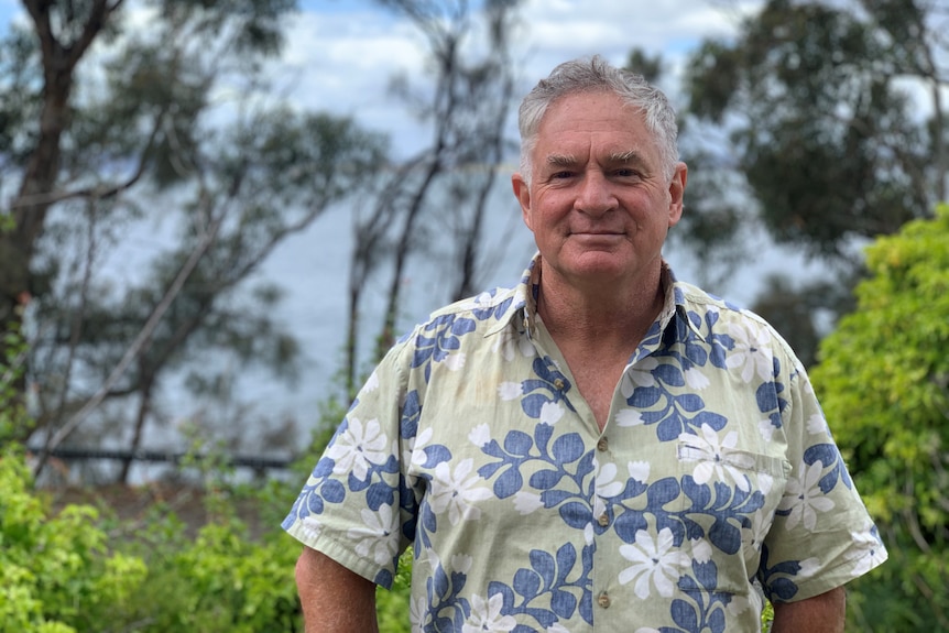 A man wearing a blue, white and green patterned shirt stands in front of a garden.