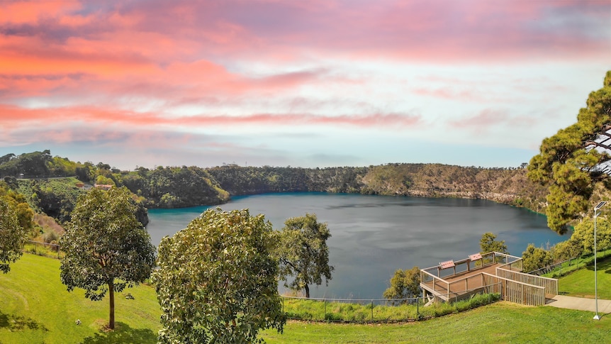 A blue lake in a crater, surrounded by green lawns and trees, with an early morning sky.