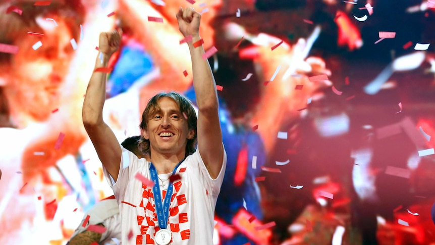 Luka Modric, wearing a silver medal, stands with his hands aloft as confetti falls around him