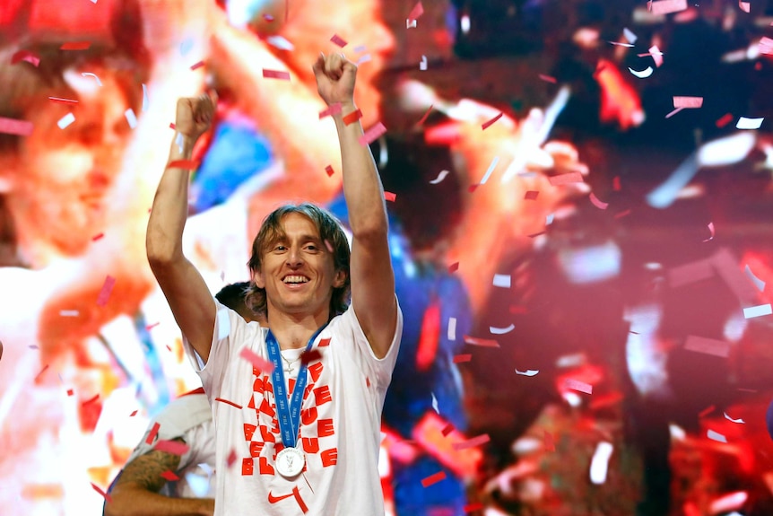 Luka Modric, wearing a silver medal, stands with his hands aloft as confetti falls around him