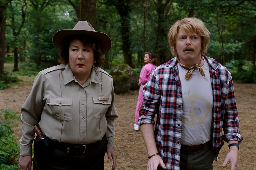White woman in a park ranger outfit stands looking trepidatious beside a white man in a white and red flannel shirt.