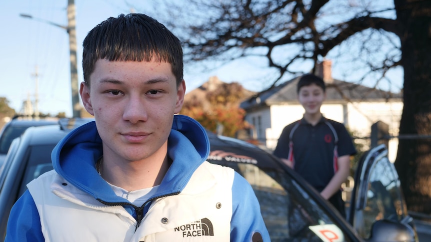 A boy smiling to camera in the foreground, a car with a P plate and a younger boy in the background.