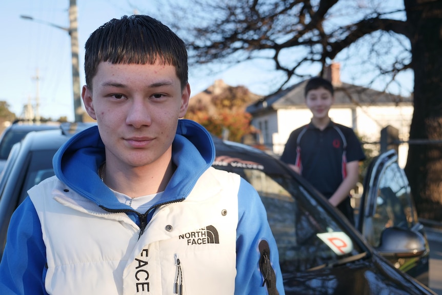A boy smiling to camera in the foreground, a car with a P plate and a younger boy in the background.
