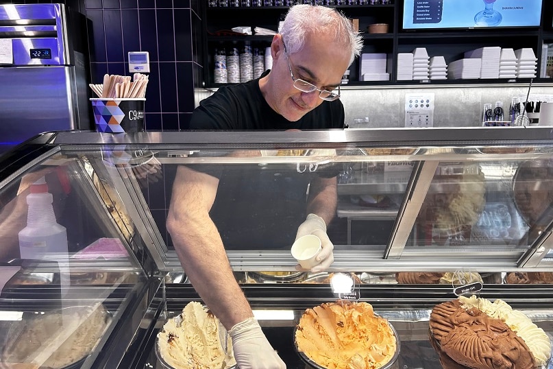 A man in a black t-shirt and glasses is reaching for an ice cream shelf to scoop ice cream into a cup.
