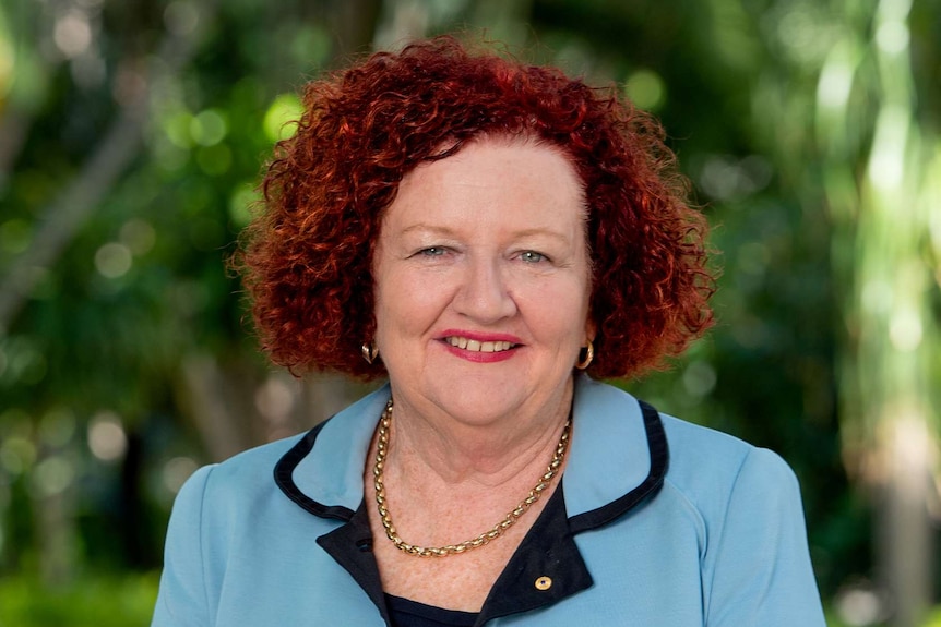 An older, red-haired woman in a light green jacket with black piping over a black dress stands near treed gardens