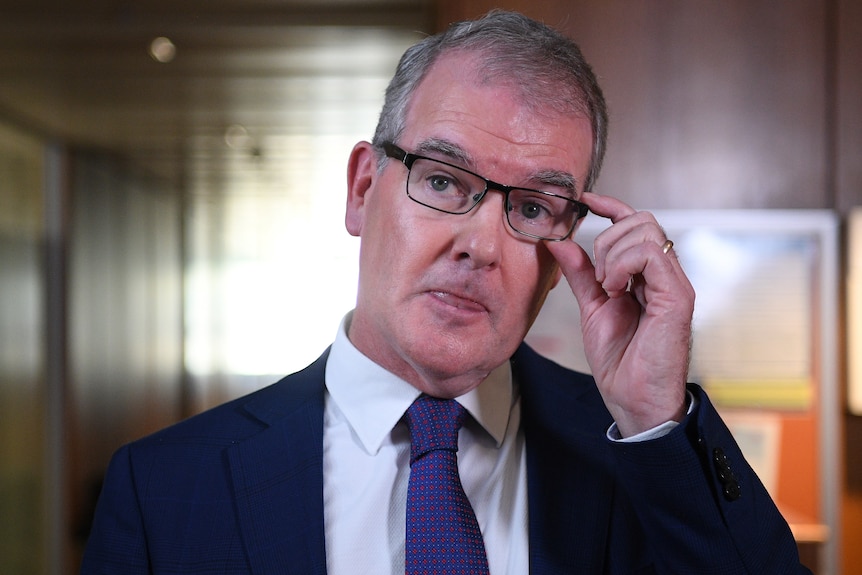 A middle-aged man in a dark suit adjusts his glasses.