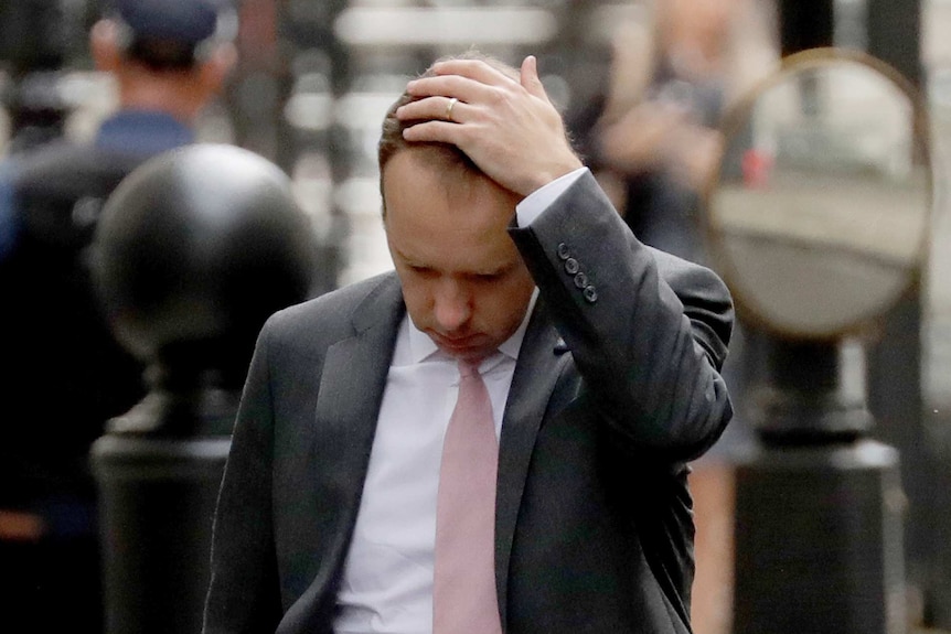 Man in grey suit touches his head, wedding ring visible