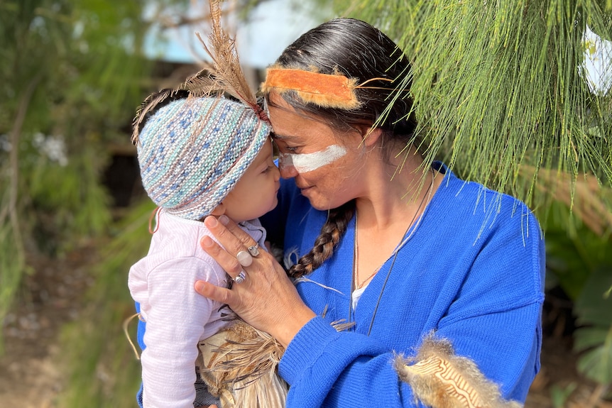 A gunditjmara mother stands holding her baby, both adorned with indigenous wears