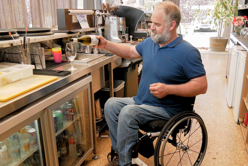 A man in a wheelchair pouring a bottle of wine behind a bar.