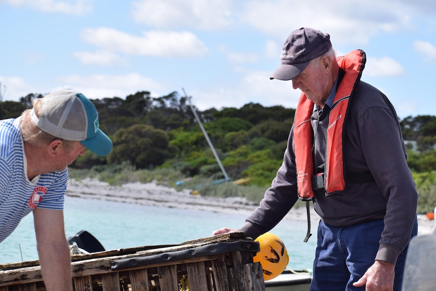 A younger man and an older man stand by a boat that's been pulled to shore. In the background is a beach and the water.