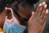 An Africa-descended woman with long hair in dreadlocks wearing pink and blue mask holds hand in front of her in prayer pose