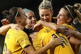 Five Matildas players embrace in celebration of a goal.