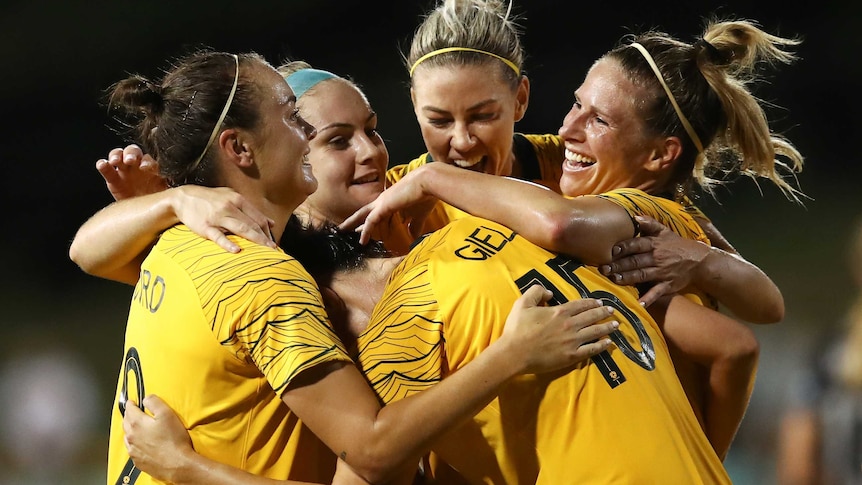 Five Matildas players embrace in celebration of a goal.