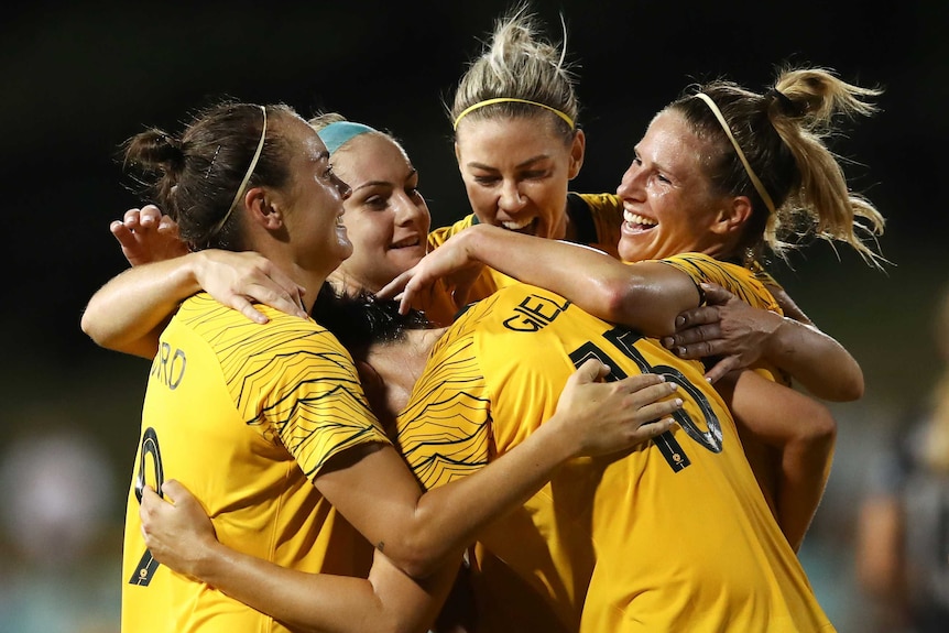 Five Matildas players embrace in celebration of a goal.