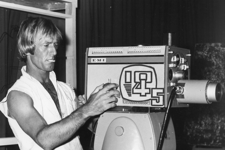 A black and white photo of actor paul hogan operating a camera, cigarette in his mouth with cut off T-shirt