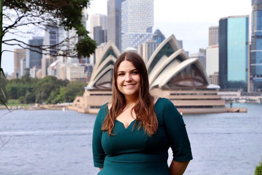 woman stands in front of the sydney opera house in green long sleeve shirt smiling with long brown hair