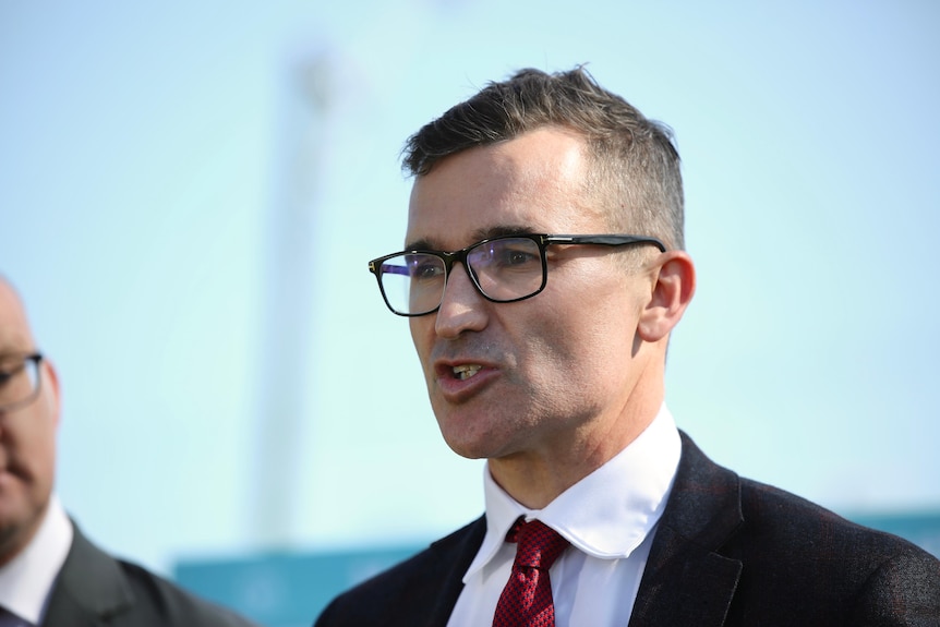 A man wearing a suit and dark-framed glasses speaks at a media conference