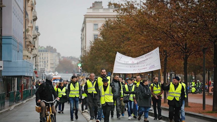 Protesters in bright yellow vests march on the street with a banner in Lyon, France, as a cyclist looks on