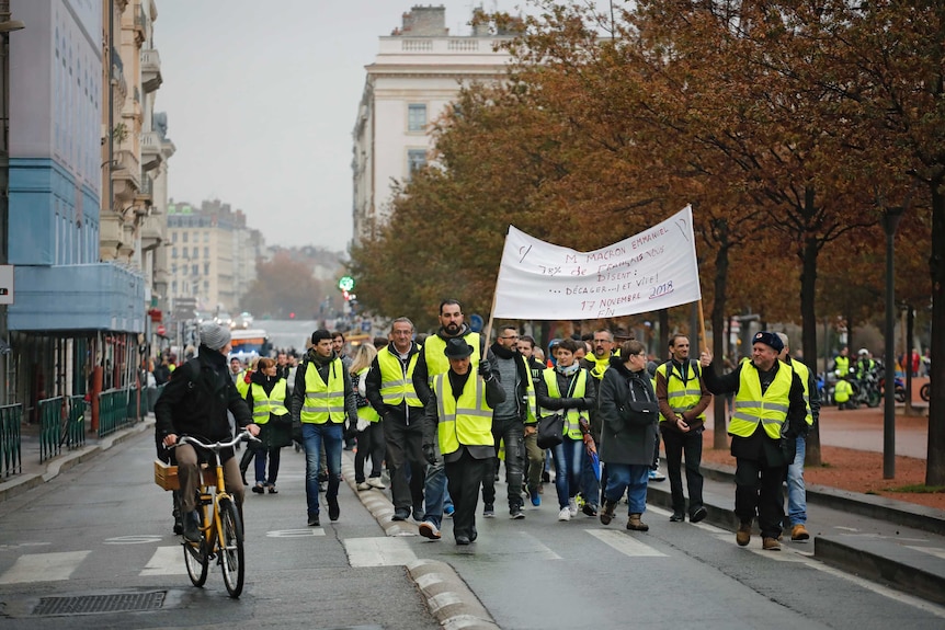 Protesters in bright yellow vests march on the street with a banner in Lyon, France, as a cyclist looks on