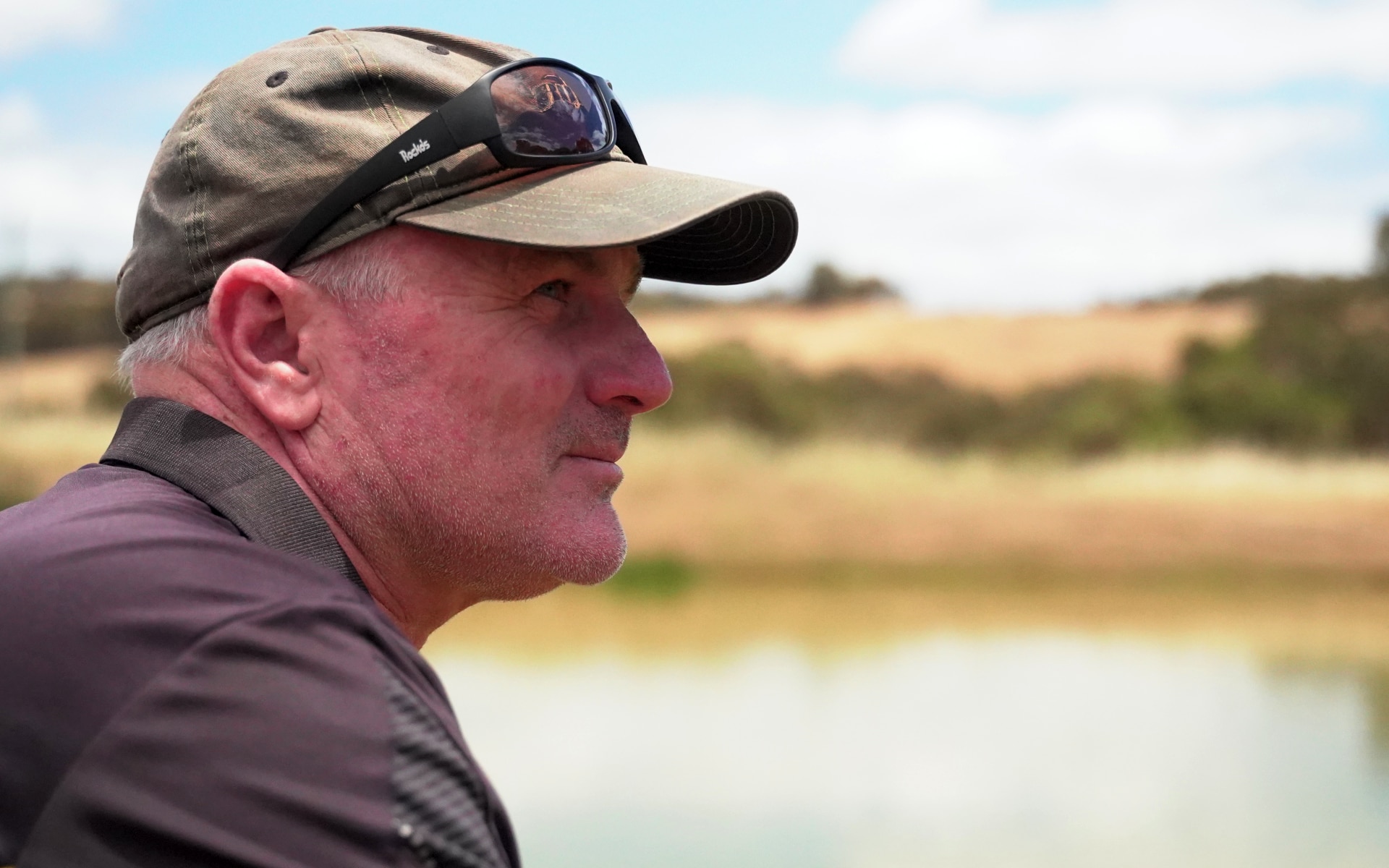 Un hombre de mediana edad se encuentra de lado frente a la cámara, se puede ver el perfil lateral de su rostro con una gorra y gafas de sol en la cabeza.