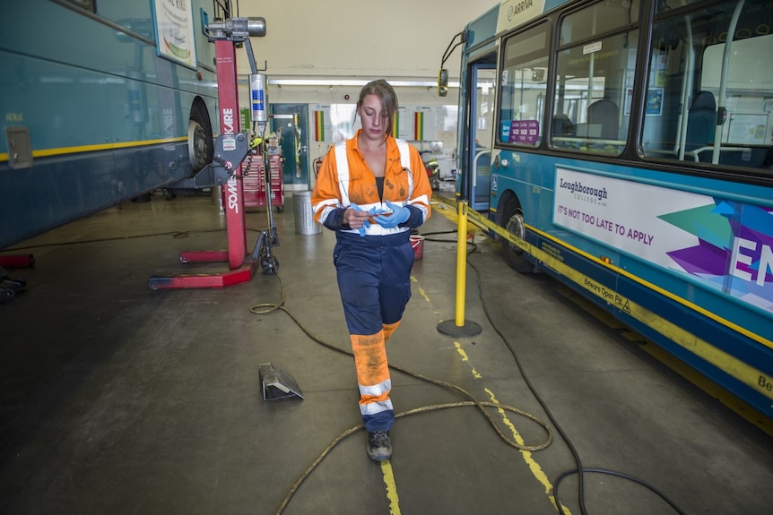 A young woman wearing high visibility workwear walks between two buses in a mechanical workshop