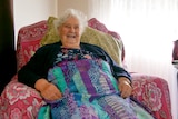 Smiling elderly woman sits in chair with knitting on her lap.