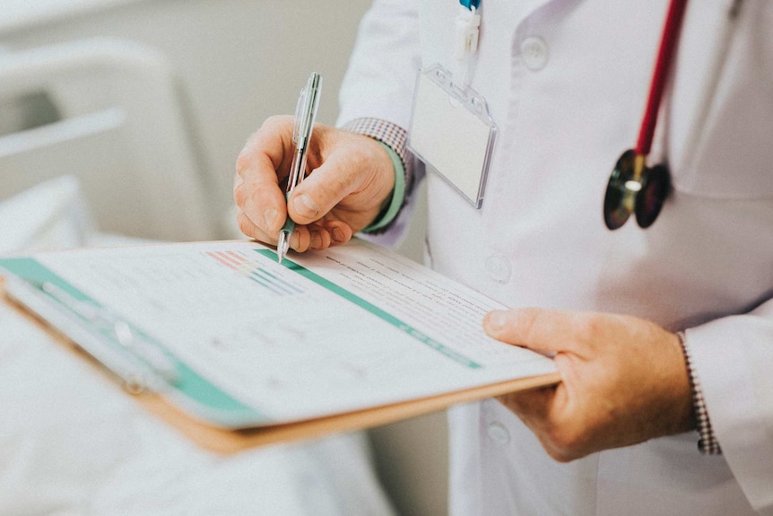 A close-up shot of a doctor noting down symptoms of a patient on a form with a stethoscope hanging around his neck.