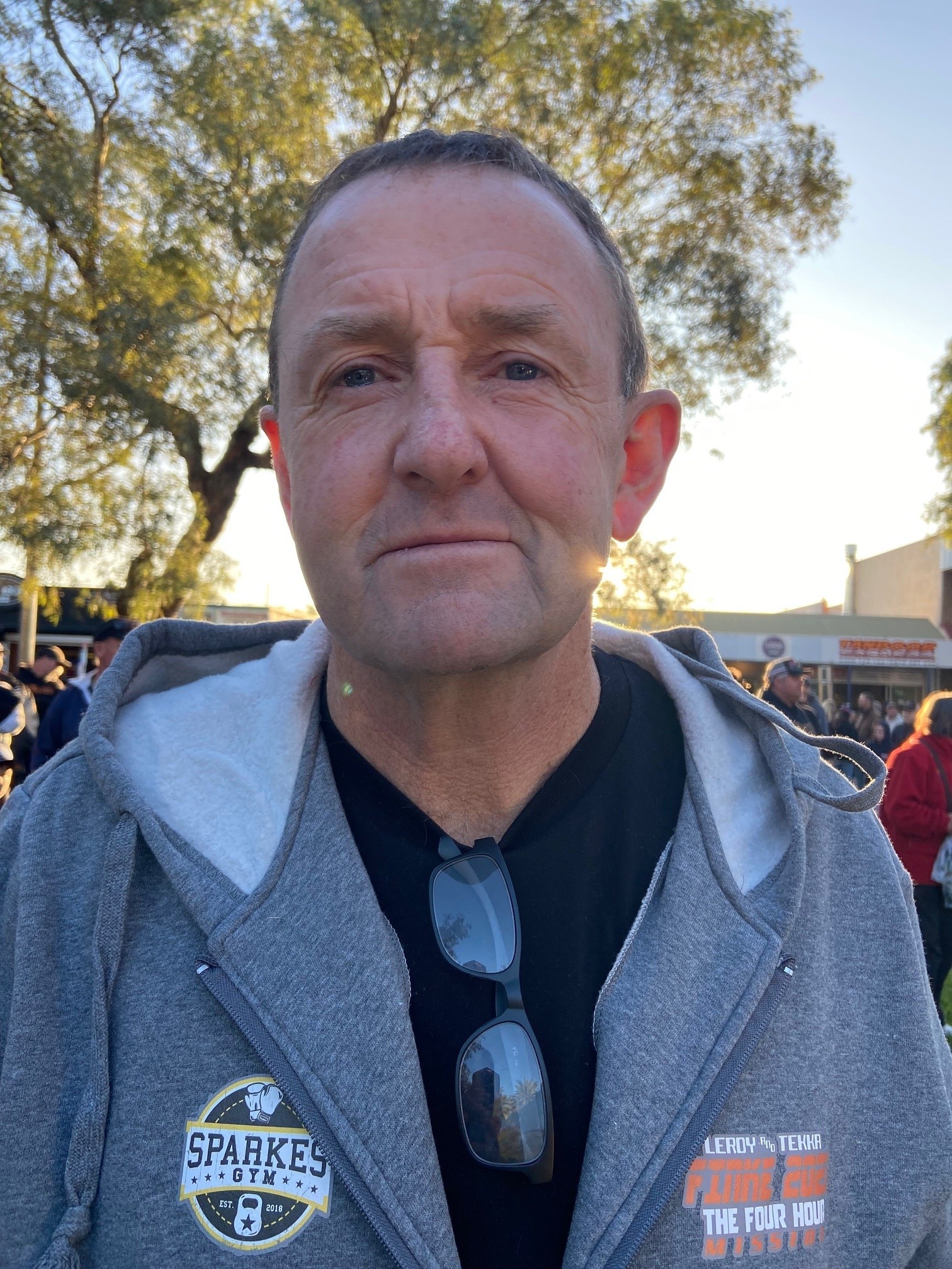 Un hombre viste una camiseta negra, una sudadera con capucha gris y gafas de sol.