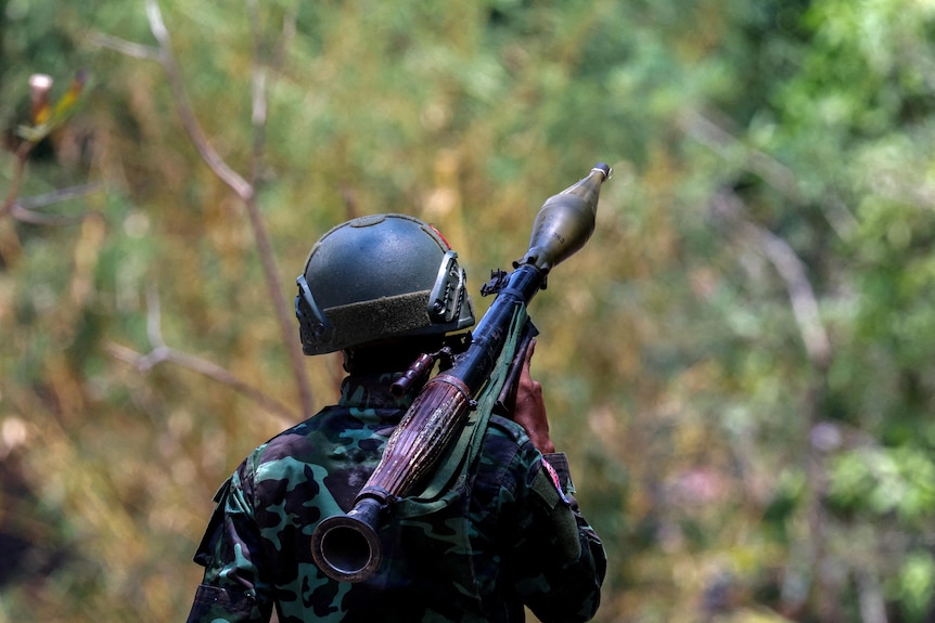 A soldier in fatigues carries a cylindrical rocket launcher on their shoulder as they walk thorugh jungle. 