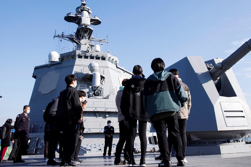 Young people in civilian clothing stand on board a Japanese military vessel in bright sunshine.