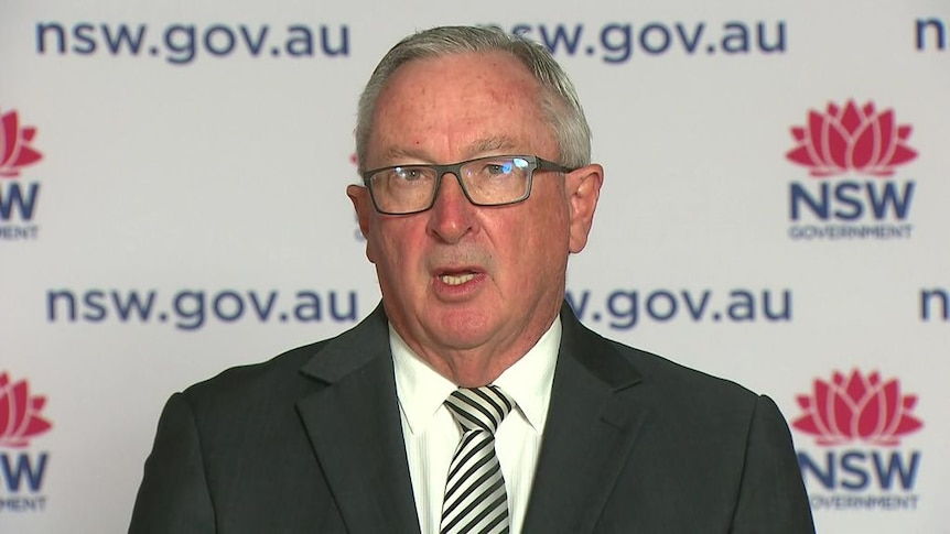 A bespectacled man in a suit, standing in front of a New South Wales Government banner.
