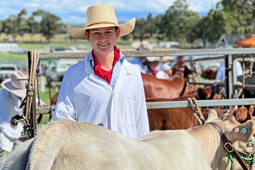 A teenage girl in a white coat and broad-brimmed hat stands behind a Charolais cow, smiling.