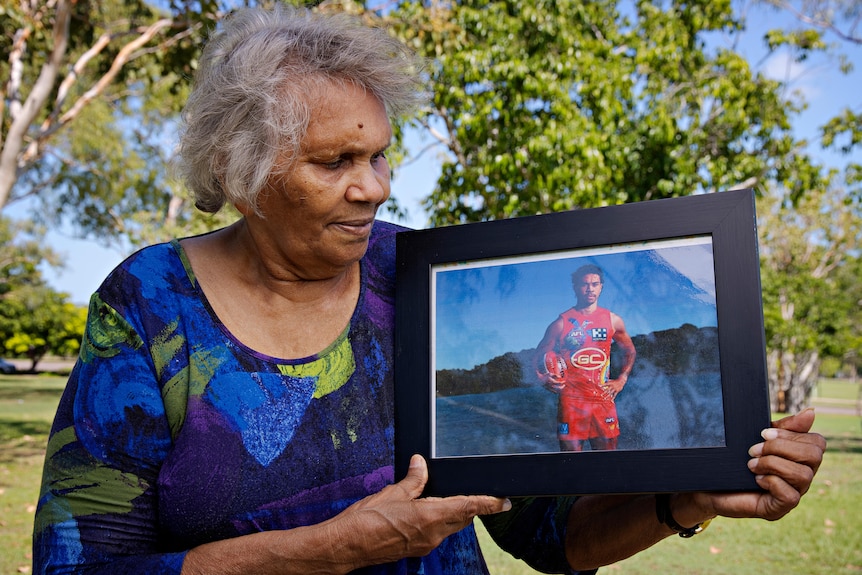 An older woman looks at a framed photo she's holding of a young man in a football jersey. 