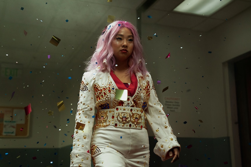 Asian-American woman with pink hair wearing Elvis-like bedazzled white suit walks through confetti-filled corridor.