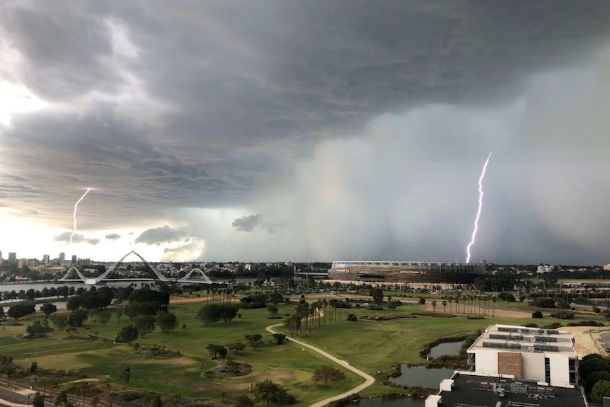 Two bright lightning bolts strike from dark storm clouds, one over Perth Stadium and the other in the distance.