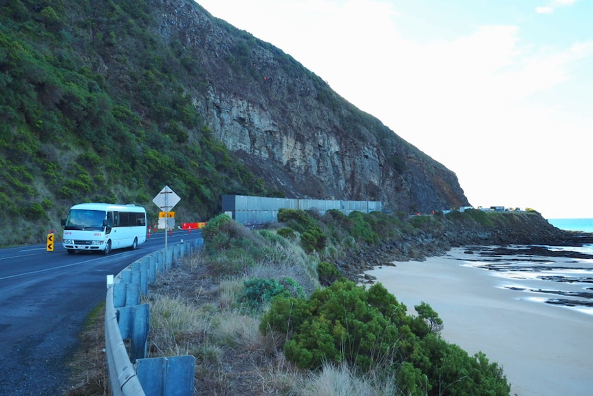 A white mini bus drives past a row of grey shipping containers which separate a coastal road from high cliffs above.