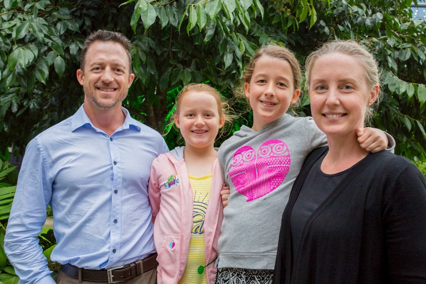 Chris wearing a blue shirt, with his two young daughters and wife Elsa, standing in front of a green bush.