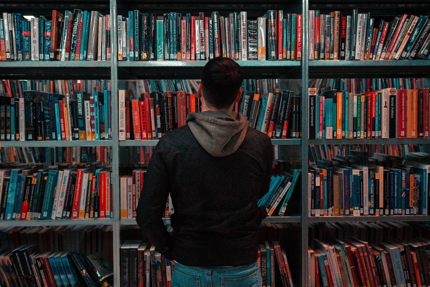 Anonymous man with a hoodie stands can be seen from behind in front of a book case in a library.